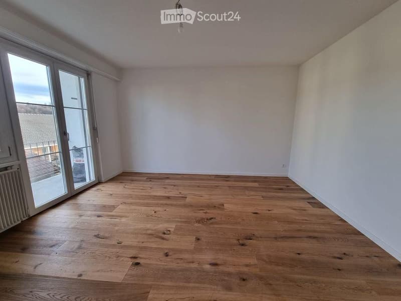 Empty room with wooden flooring, white walls, large glass sliding doors leading to a balcony, and a radiator on the left side.