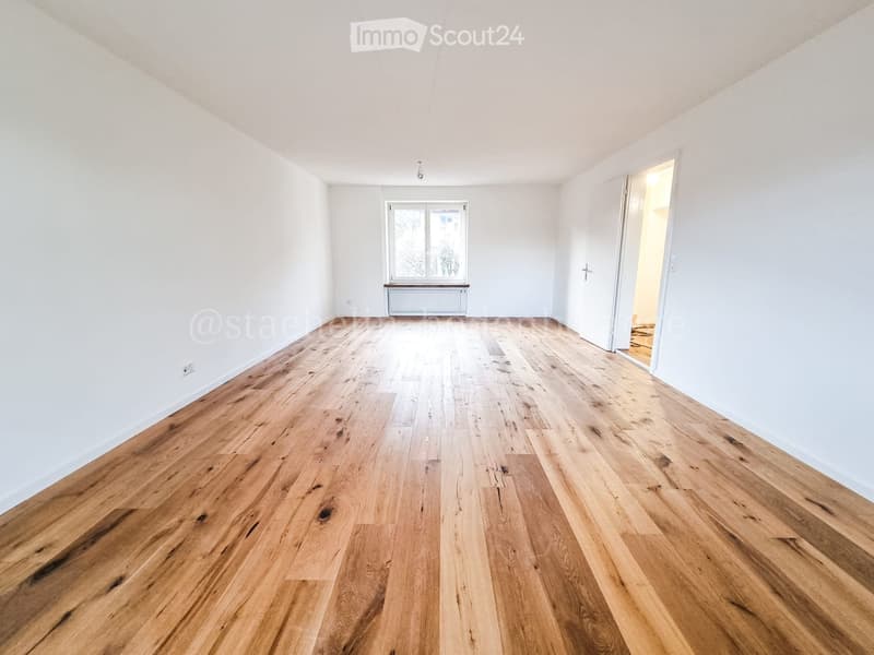 Empty room with wooden flooring, white walls, and a window with a radiator below it. The room has a door leading to another space.