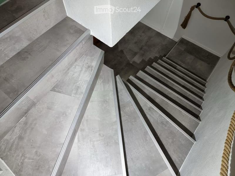 Modern interior staircase with grey concrete steps and white walls, featuring a rope handrail on one side.