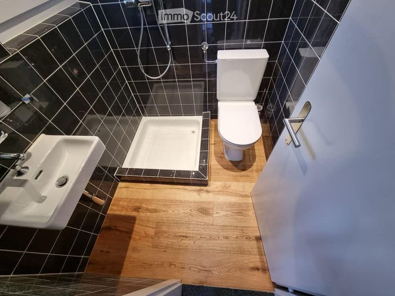 Modern bathroom with dark tiled walls, light wooden floor, toilet, sink, and shower stall.