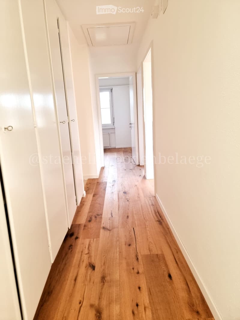 A long, narrow hallway with light wooden flooring and white walls, featuring several closed doors on both sides and a visible ceiling vent.
