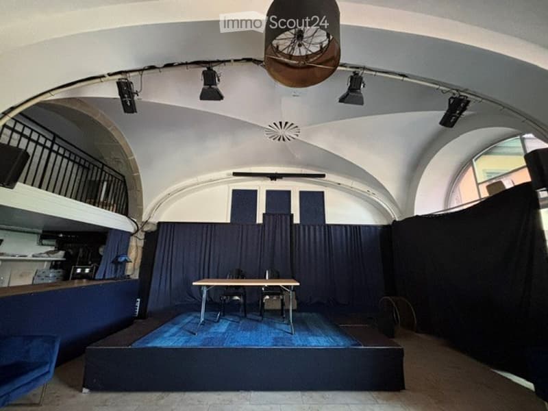 A small, modern room with a raised platform, a table, two chairs, and dark blue curtains. The ceiling features industrial-style lighting fixtures and a ventilation system.