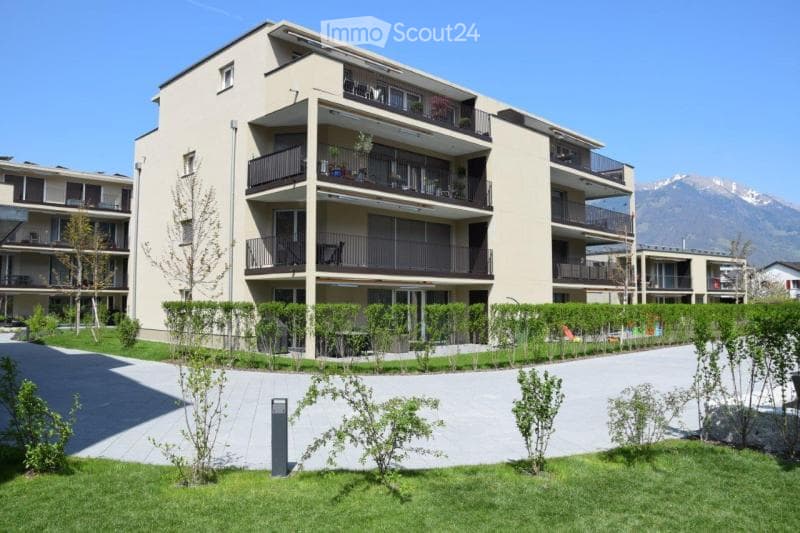 Modern multi-story apartment building with balconies, surrounded by greenery and a clear view of mountains in the background.