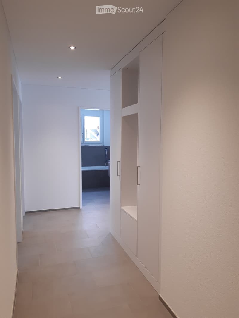 Modern hallway with white walls, light-colored tiled floor, built-in white storage units, and recessed ceiling lighting. An open doorway leads to another room.
