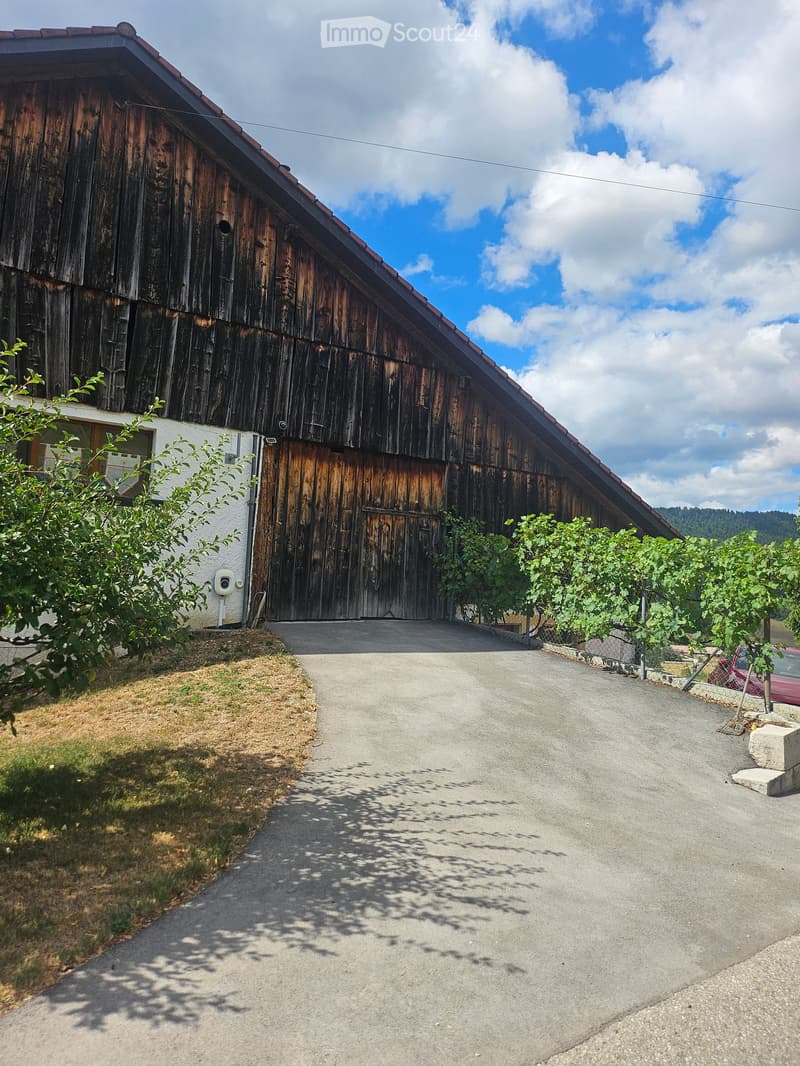 A large wooden barn with a sloped roof and a concrete driveway leading to it, surrounded by greenery and a small white building.
