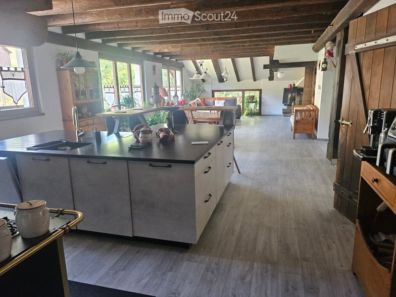 Open-plan kitchen and living area with wooden beams, grey cabinets, island, sink, coffee maker, and dining area with table and chairs.