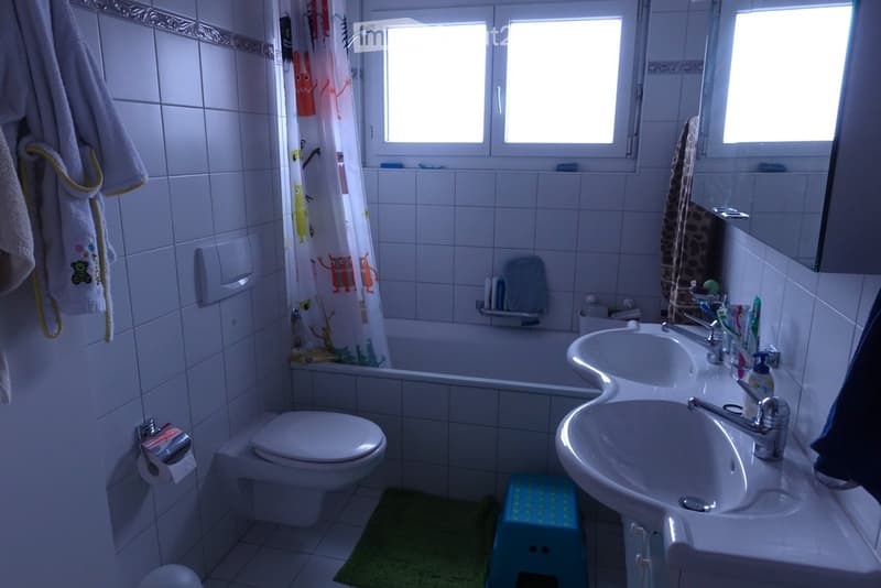 Bathroom with white tiles, a toilet, a double sink with two faucets, a bathtub with a colorful shower curtain, a towel hanging on the wall, and a small green rug on the floor.