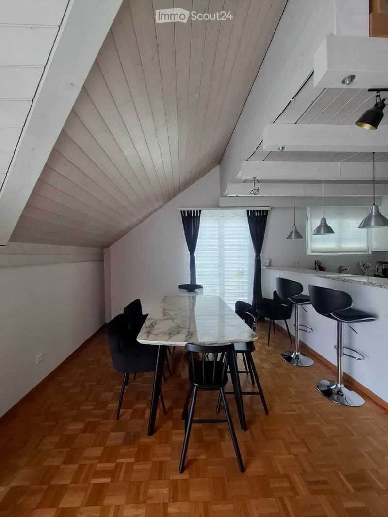 Modern attic room with wooden flooring, slanted wooden ceiling, glass dining table with marble top, black chairs, and a kitchen area with bar stools.