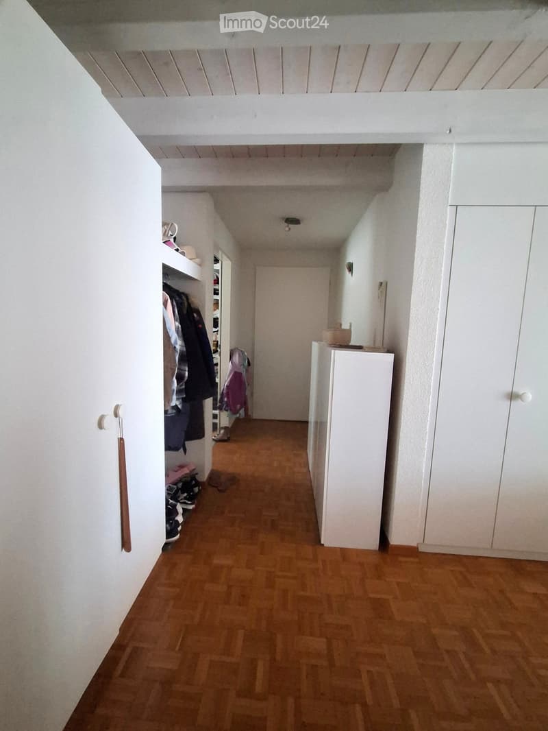 A narrow hallway with wooden flooring, white walls, and built-in storage units on both sides. Clothes and shoes are visible in the left storage area, and a small table with items sits on the right.