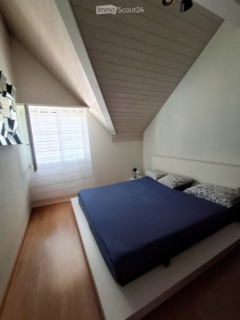 Cozy attic bedroom with a raised wooden bed, blue bedding, and a nightstand with a lamp. The room features wooden flooring and a slanted ceiling with exposed beams.
