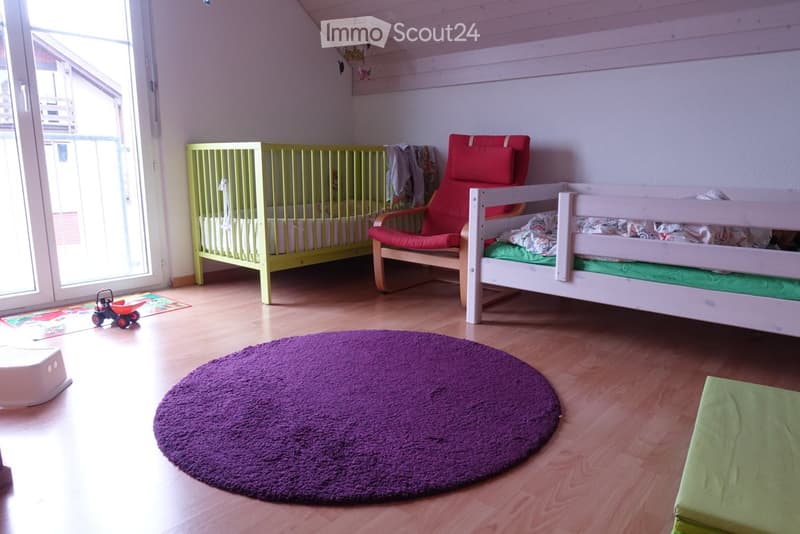 Brightly lit children's bedroom with wooden flooring, a green crib, a white daybed, a red chair, a purple round rug, and a small toy car on the floor.