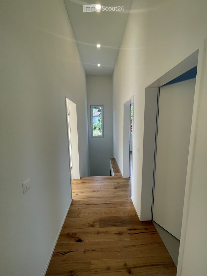 Empty hallway with wooden flooring, white walls, and recessed ceiling lighting. Features a small window at the end and several open doors leading to other rooms.