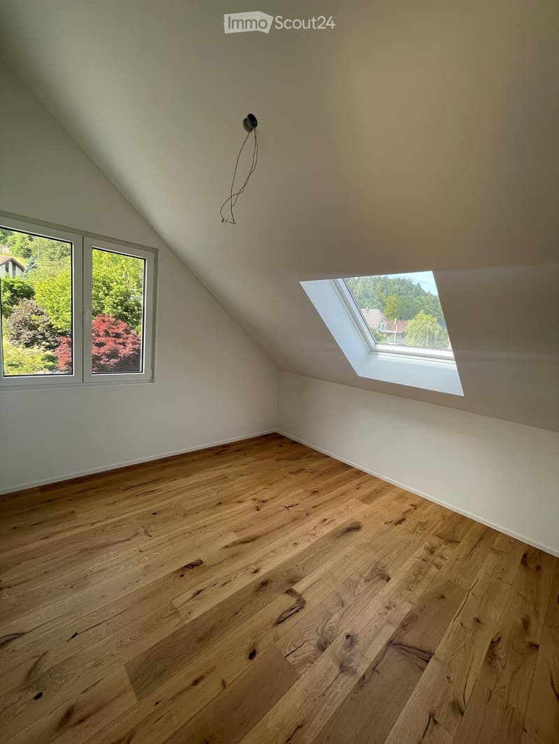 Empty attic room with wooden flooring, white walls, and large windows offering natural light and a view of greenery outside.