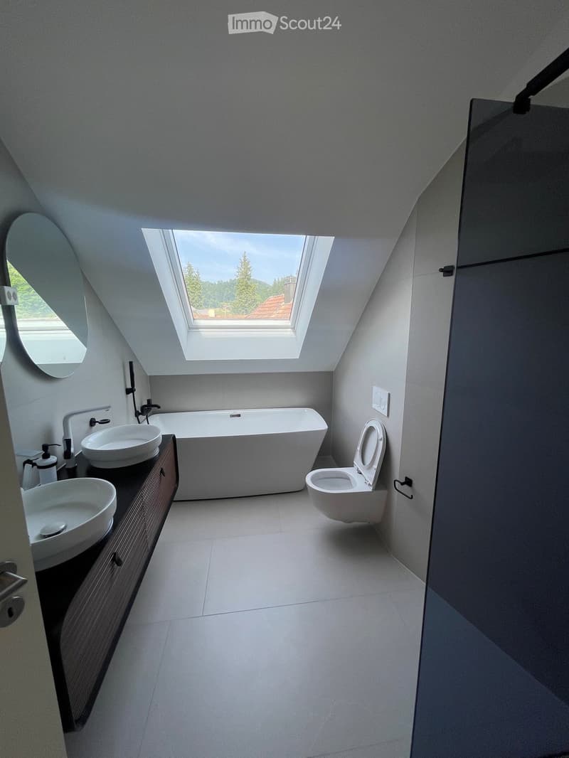 Modern bathroom with a large skylight, white bathtub, two sinks, toilet, and a round mirror.