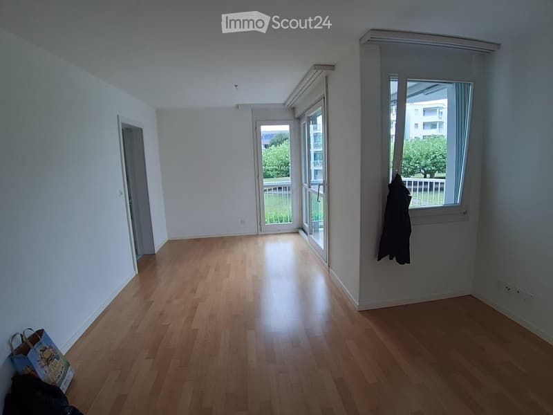 Empty room with wooden flooring, white walls, and large windows. A coat is hanging on the wall, and a shopping bag is placed near the entrance.