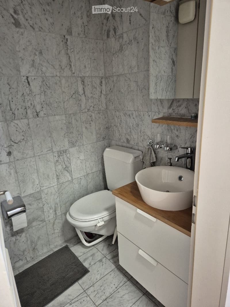 Modern bathroom with marble tiles, white toilet, round sink on wooden countertop, mirror, and towel.