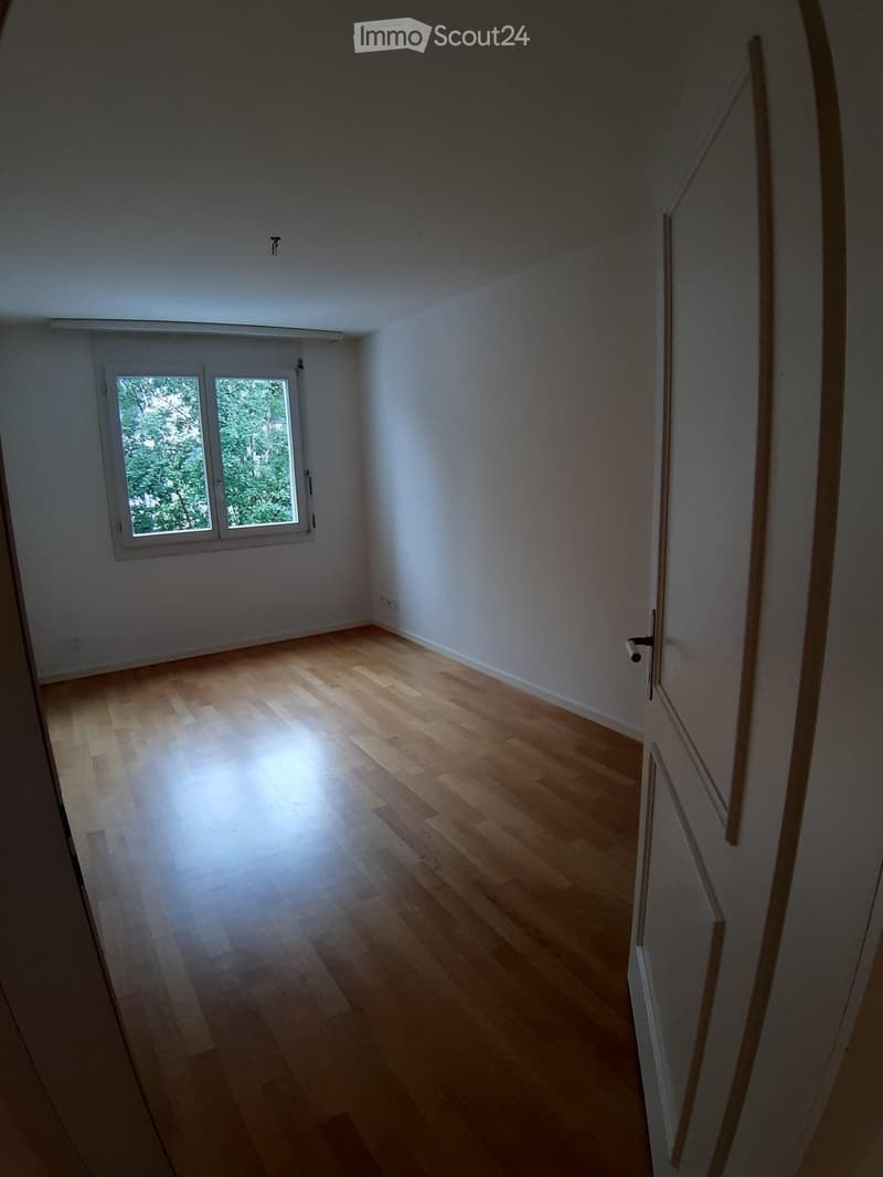 Empty room with light wooden flooring and white walls, featuring a window with white frames and a door with a silver handle.