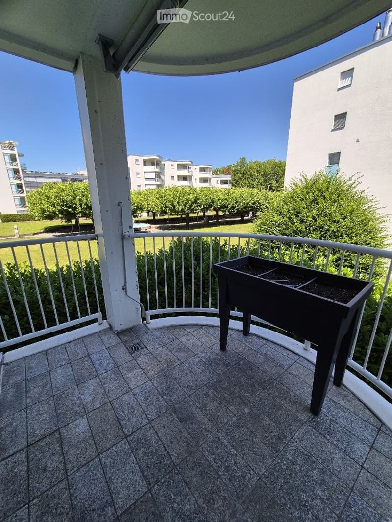 Balcony with tiled floor, white metal railing, and a small black planter. The view shows a park with trees and buildings in the background.