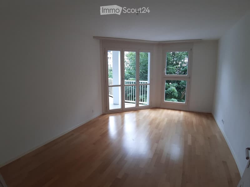Empty room with light wooden flooring and large windows, including a sliding glass door leading to a balcony.
