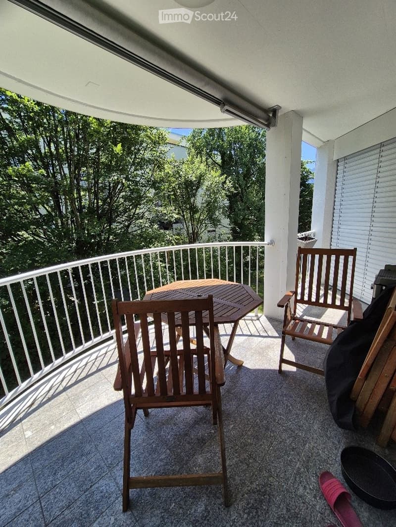 Balcony with wooden table and chairs, overlooking a green area with trees.