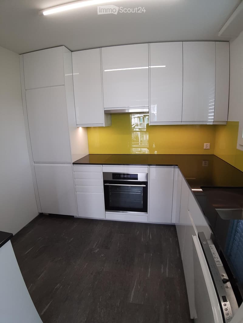 Modern kitchen with white cabinets, dark countertops, and a built-in oven. The backsplash is a bright yellow, and the floor is dark wood.