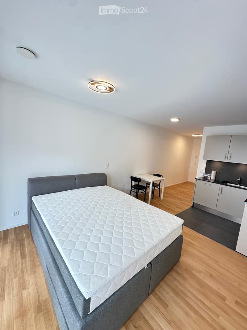 a large bed with white mattress and gray frame, white walls, dining table, black chairs, wooden floor, kitchen with sink, cabinets, countertop