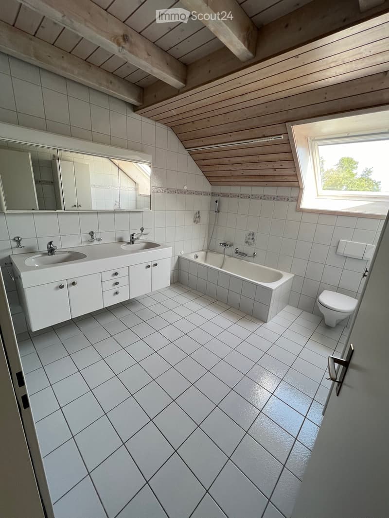 Modern bathroom with white tiles, double sink, bathtub, toilet, and large mirror. Features a skylight window and wooden slanted ceiling.