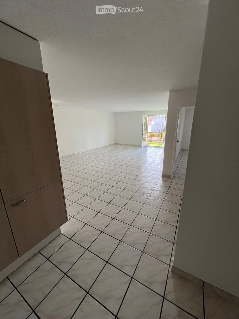 Empty hallway with tiled floor and white walls, leading to an open door that reveals a garden area outside.