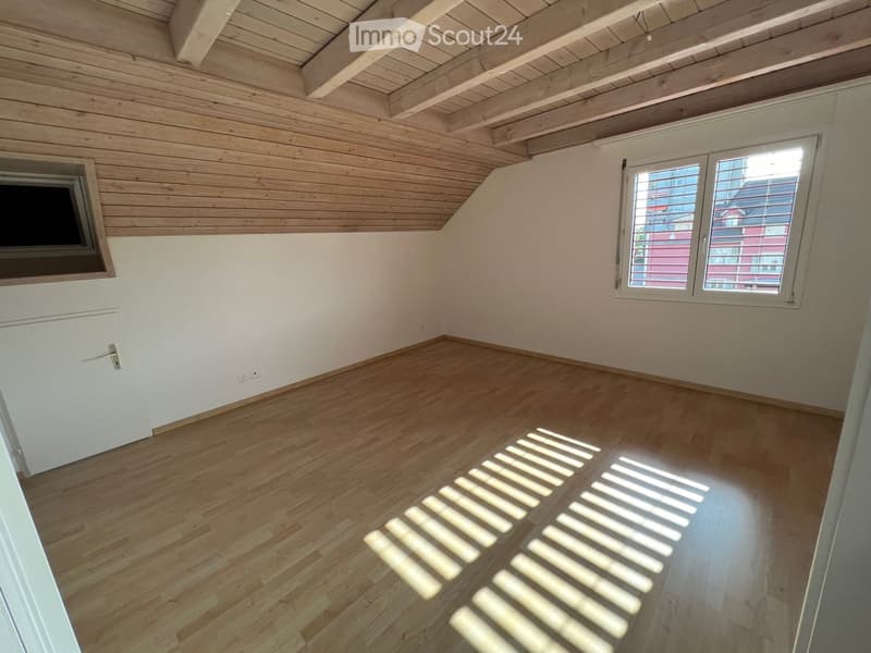 Empty room with wooden flooring and exposed wooden ceiling beams. Two windows provide natural light, one with blinds partially drawn.