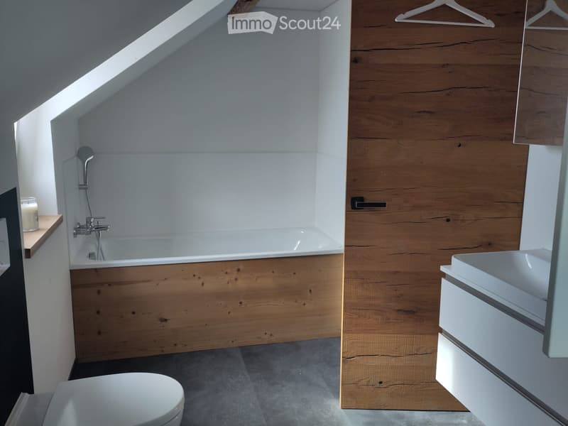 Modern bathroom with a wooden accent wall, bathtub, toilet, sink, and a small shelf holding a candle. The space features a skylight window and sleek fixtures.