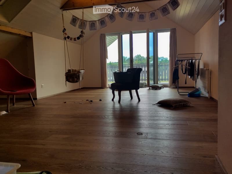 A cozy, slanted-ceiling room with wooden flooring, a red chair, a hanging swing, a small black chair, a clothing rack, and a pillow on the floor.
