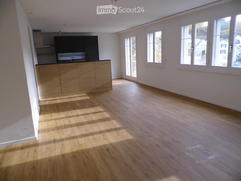 Empty room with wooden flooring, white walls, and large windows allowing natural light. A small kitchen area with cabinets is visible in the background.