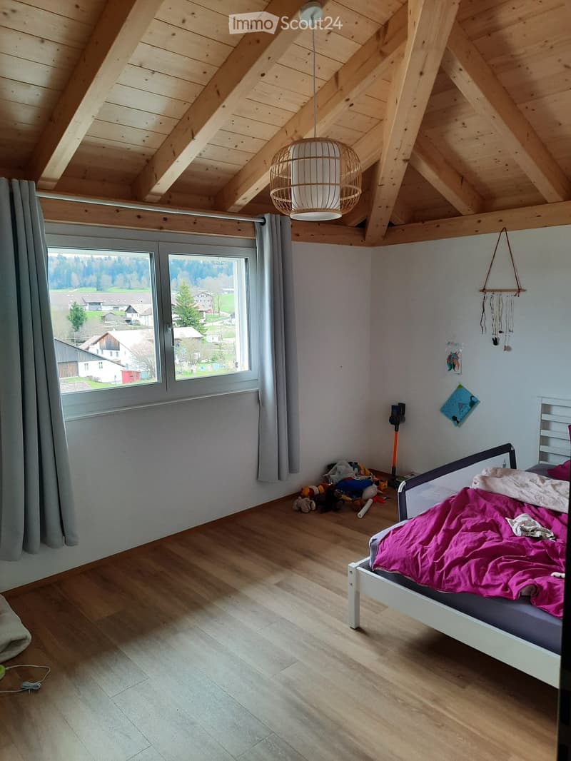 Bright bedroom with wooden slanted ceiling, large window with view, bed with pink blanket, toys on floor, wicker pendant light, and decorative items on walls.
