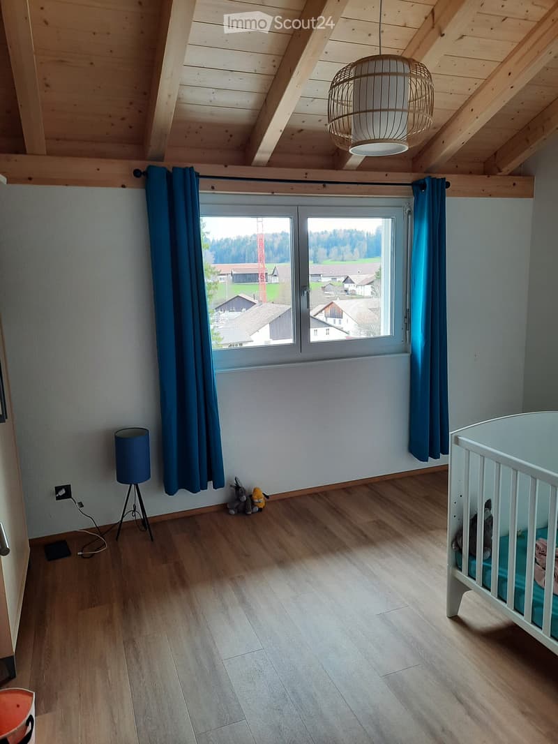 Bright, airy bedroom with wooden floors, large window with blue curtains, a crib, a lamp, and toys.