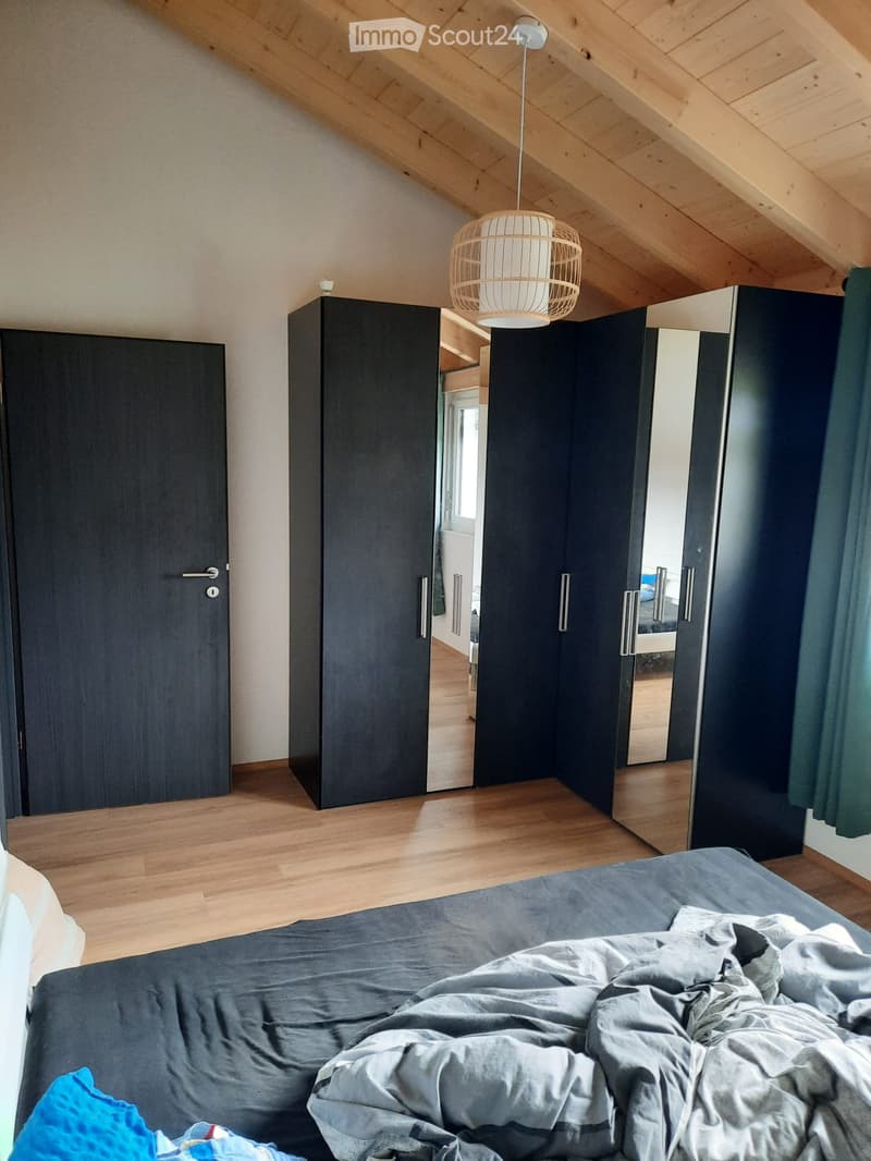 Modern bedroom with wooden floor, slanted wooden ceiling, grey bedding, dark wardrobes, and a hanging woven pendant light.