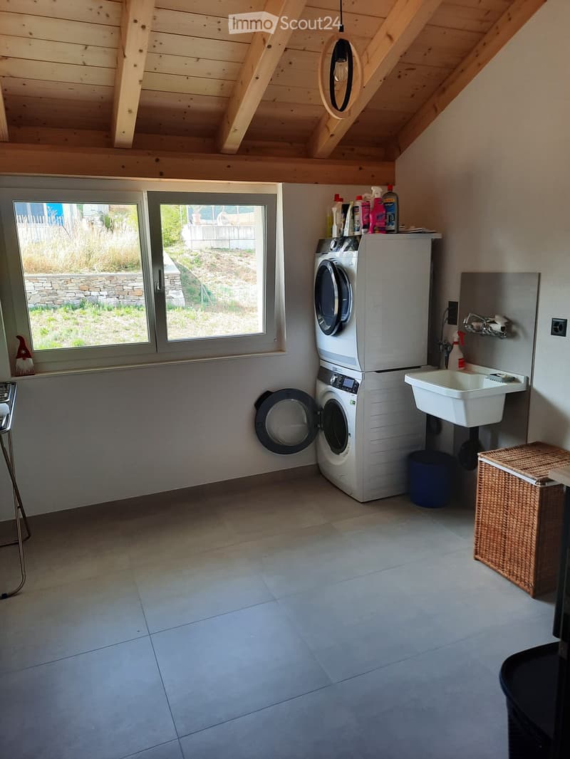 Well-lit laundry room with a washing machine, dryer, refrigerator, sink, and cleaning supplies on top. Features a wicker basket and a small stool near the window.