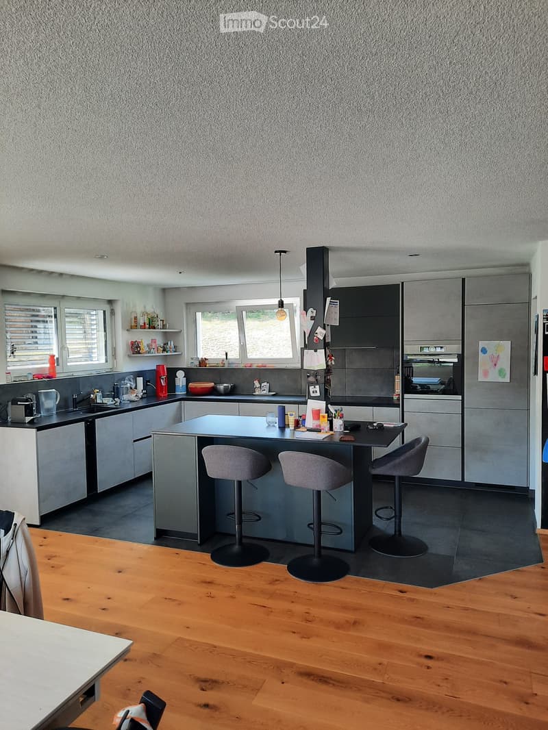 Modern kitchen with grey cabinets, central island, bar stools, and various appliances including a fridge, oven, and coffee maker. The space features wooden flooring and large windows.