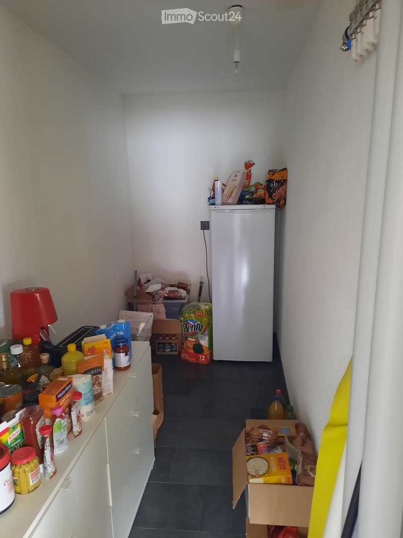 A small kitchen with white cabinets, a refrigerator, a toaster, and various food items and supplies on the counter and floor.