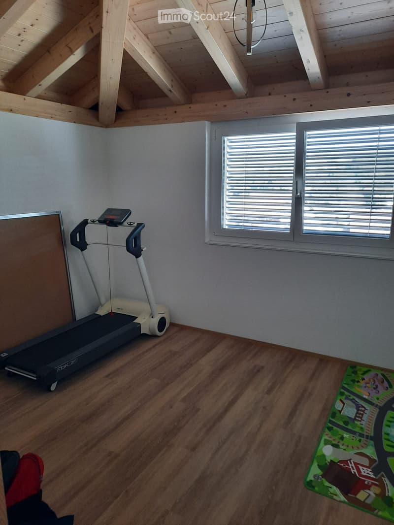 Exercise room with wooden floor, exposed wooden ceiling beams, large window with blinds, treadmill, and play mat.