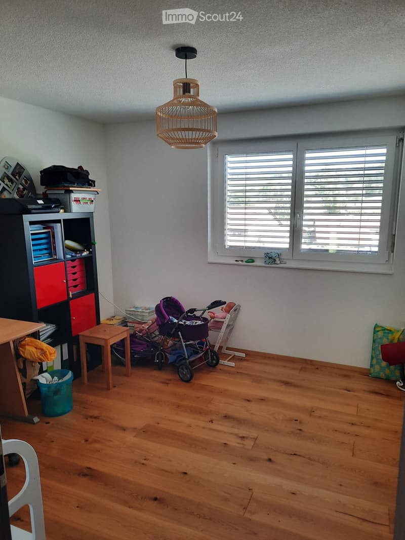 A well-lit room with wooden flooring, a large window with blinds, a hanging woven lamp, a small wooden table, a stool, a baby stroller, a baby chair, a bookshelf, and a trash bin.