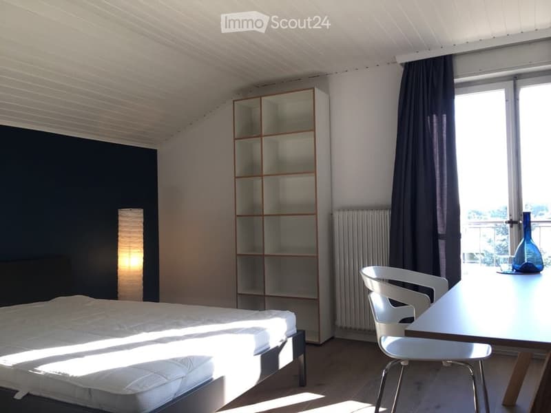 Modern bedroom with a white bed, a white chair, a small table, a shelf unit, and a blue vase on the table. The room has large windows with dark curtains and a view outside.