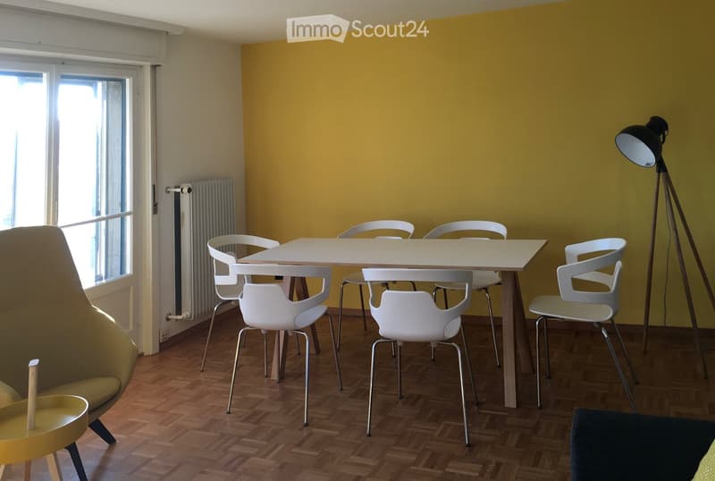 Bright room with wooden flooring, a yellow accent wall, and modern furniture including a white table, six chairs, a yellow stool, and a floor lamp.