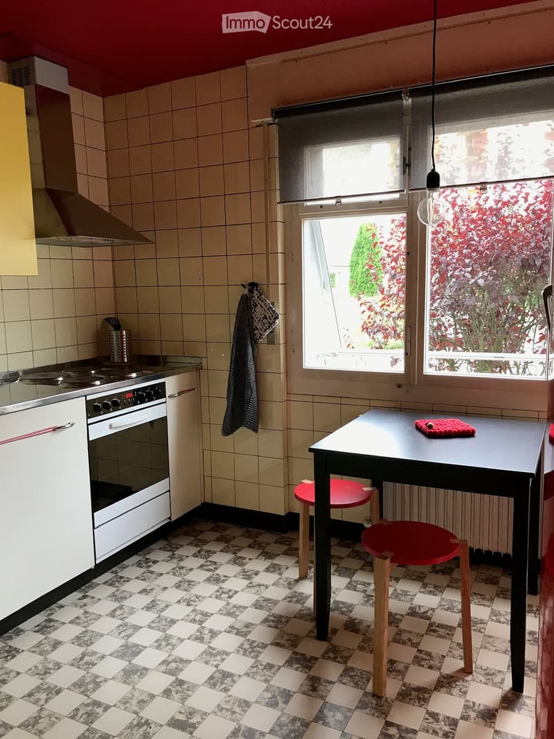 A small kitchen with tiled walls, a checkered floor, and a window overlooking a garden. Features include a white oven, a black table with red chairs, and a hanging towel.