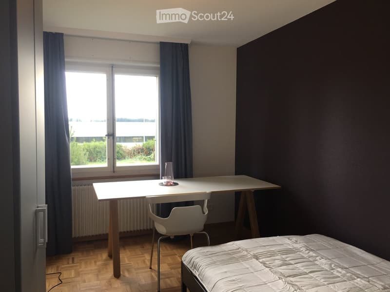 A small, furnished room with a bed, a white table, a chair, and a cup on the table. The room has wooden flooring, dark walls, and large windows with blue curtains.
