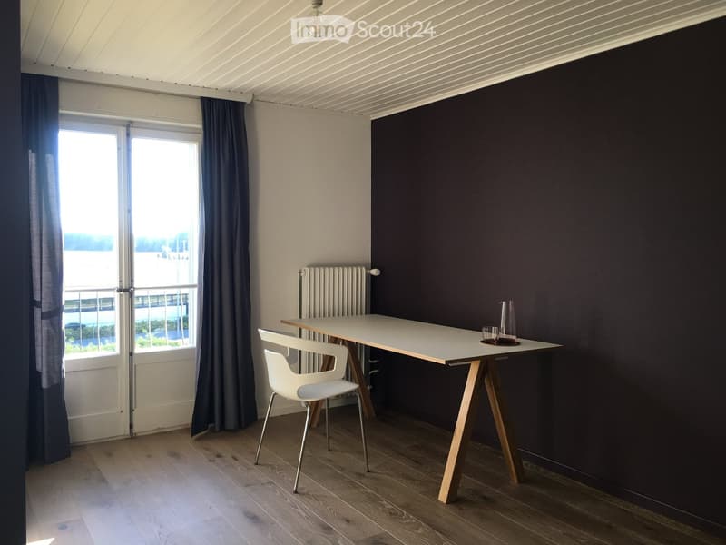 Modern, minimalist room with wooden flooring, white table, white chair, radiator, dark curtains, and a view through a large window.