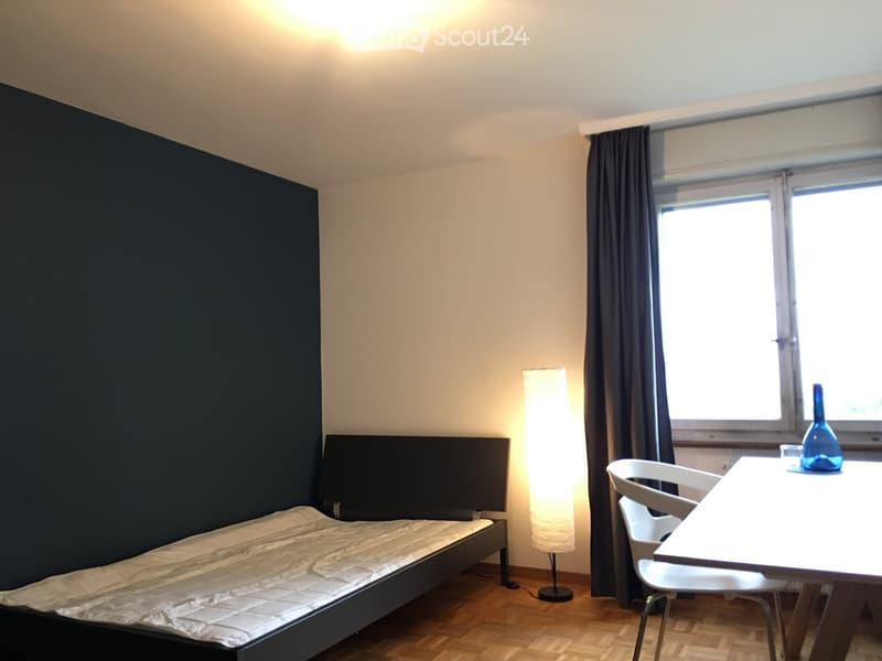 A small, modern bedroom with a single bed, a white table with chairs, a floor lamp, and a vase on the table. The room has wooden flooring and large windows with dark curtains.