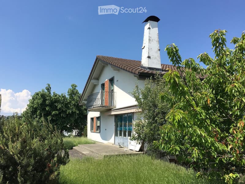 Two-story white house with a tiled roof, chimney, balcony, and surrounding greenery.
