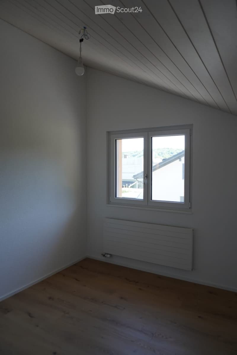 Empty attic room with wooden flooring, white walls, a single window, and a radiator. The ceiling has exposed wooden beams.