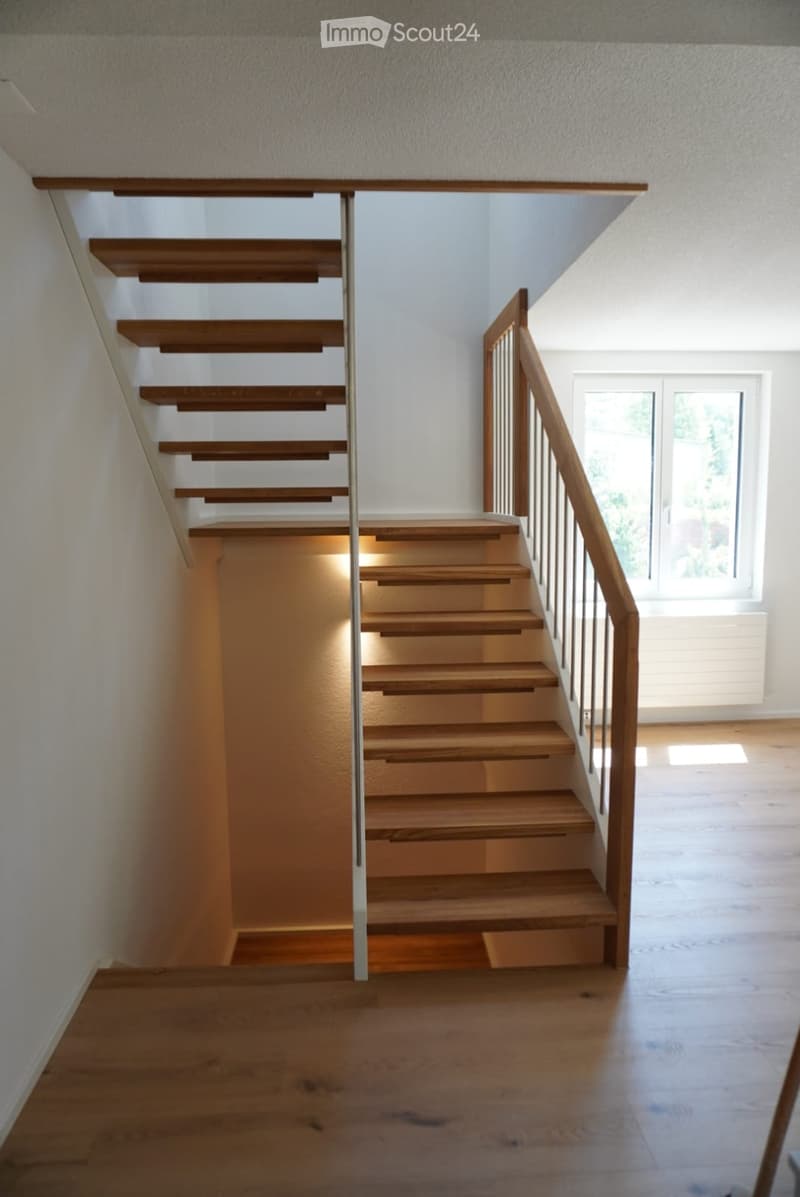 Modern wooden staircase with metal railing, illuminated steps, and a window at the end of the hallway.