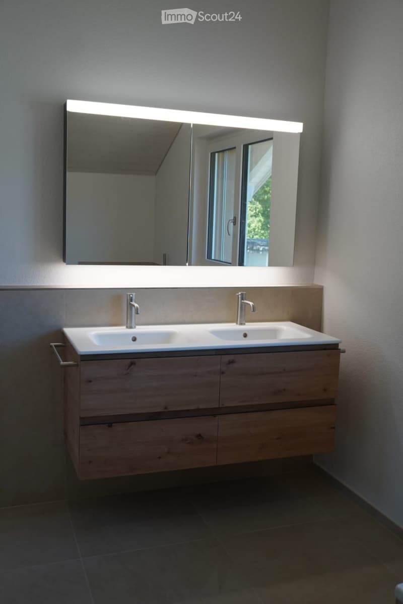 Modern bathroom with a double sink vanity, sleek wooden cabinetry, and a large mirror with integrated LED lighting. The room features a contemporary design with a neutral color palette and dark tiled flooring.